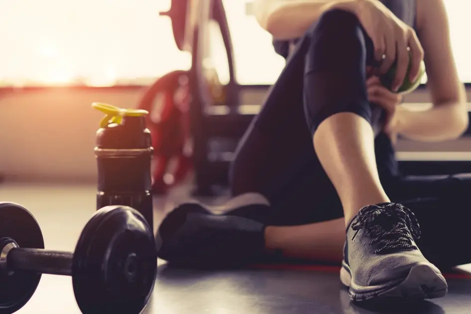 Dumbbell and water bottle on a gym floor, with a woman resting in warm, hazy sunlight after her workout with Healthwise Protein Shakes in Glendale, AZ.