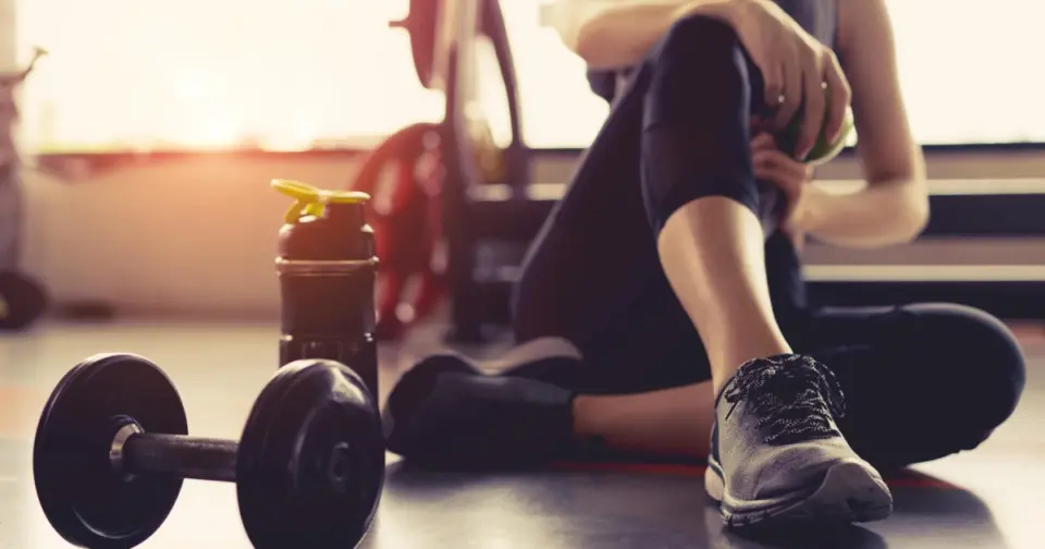 Dumbbell and water bottle on a gym floor, with a woman resting in warm, hazy sunlight after her workout with Healthwise Protein Shakes in Glendale, AZ.